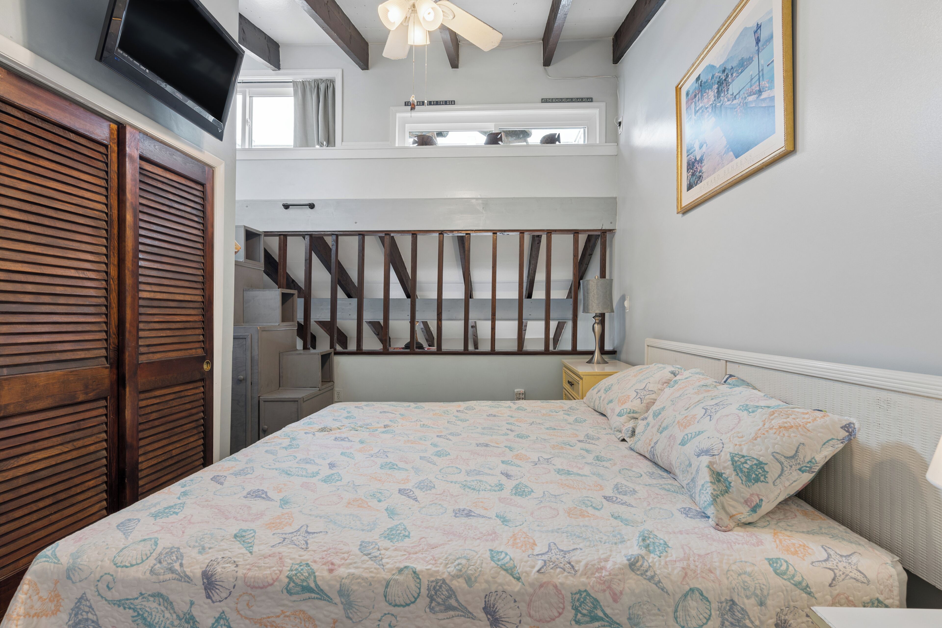 A bedroom with a bed, a TV mounted on the wall, a painting, and a staircase.