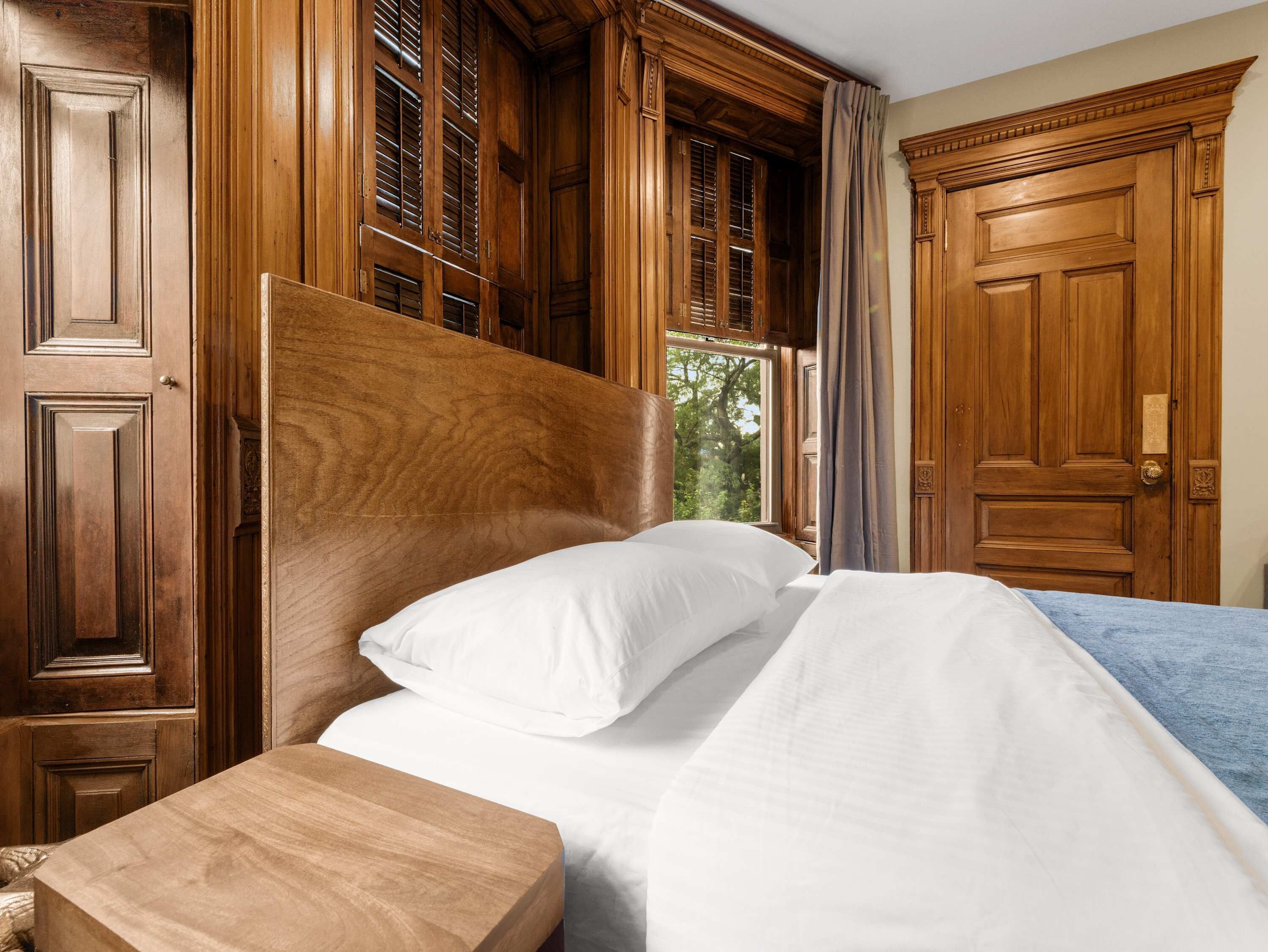 Una cama con estructura de madera y ropa de cama blanca, una mesita de noche de madera, y vistas al exterior a través de una ventana con contraventanas de madera.