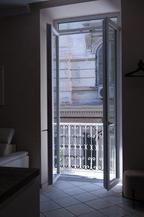 Eine offene Tür, die auf einen Balkon mit Metallgeländer, einem weißen Stuhl und einem gefliesten Boden führt.