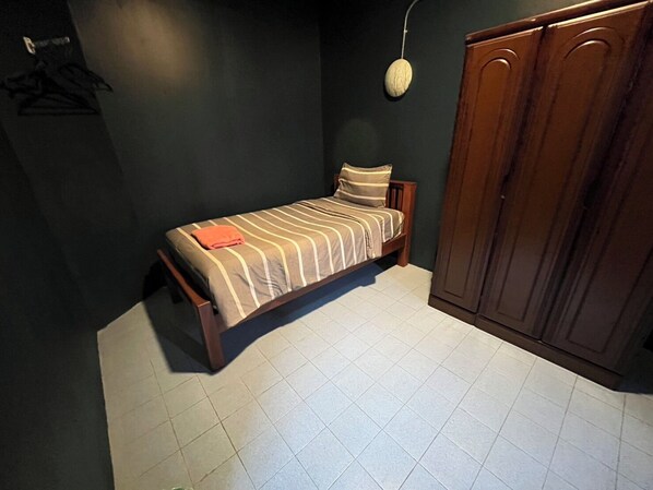 A small, single-bed room with a wooden bed frame, a striped bedspread, and a single pillow. There is a wardrobe with a dark finish to the right and a wall-mounted light fixture above the bed.