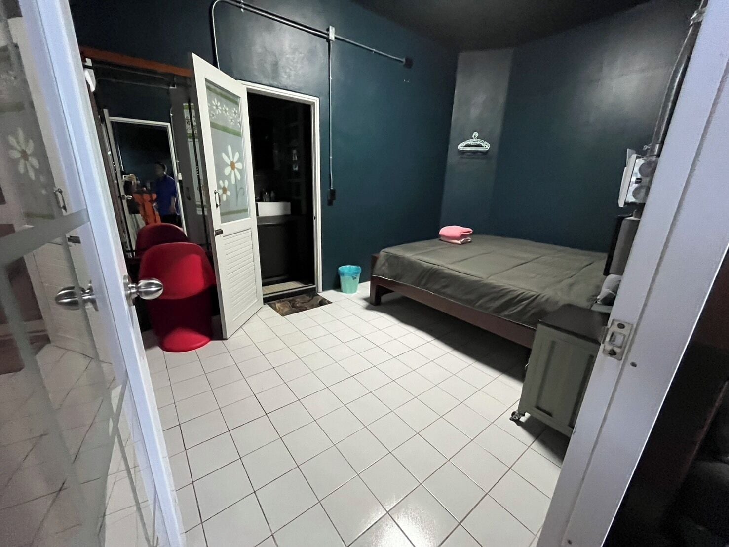 Une pièce comprenant un lit, une chaise rouge, une petite table, une poubelle bleue et une porte donnant sur une autre pièce.