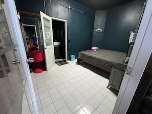 A room with a bed, a red chair, a small table, a blue trash bin, and a door leading to another room.