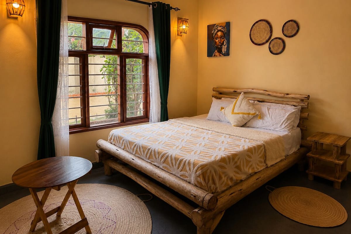 A room with a bed, a small table, a window with curtains, wall art, and a wall-mounted light fixture.