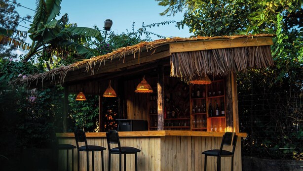 A wooden bar with a thatched roof, surrounded by greenery and equipped with hanging lights.