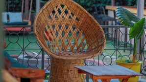 A wicker chair, a small wooden table, and a hanging light fixture in an outdoor setting with greenery.