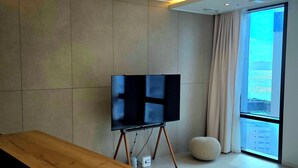 Een moderne woonkamer met een grote flatscreen televisie op een standaard, een houten salontafel, een rond poef en een groot raam met uitzicht op de stad.