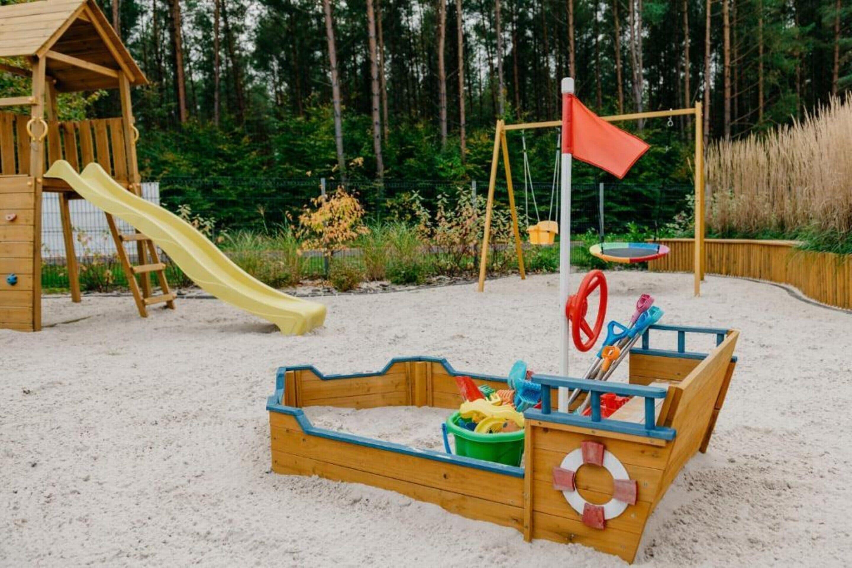 A wooden play structure with a slide and swings, surrounded by sand and greenery.