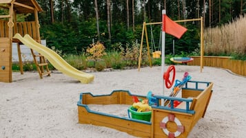 A wooden play structure with a slide and swings, surrounded by sand and greenery.