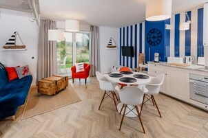 Una sala de estar moderna con un sofá azul, una silla roja, un cofre de madera y una zona de comedor con una mesa y sillas blancas.