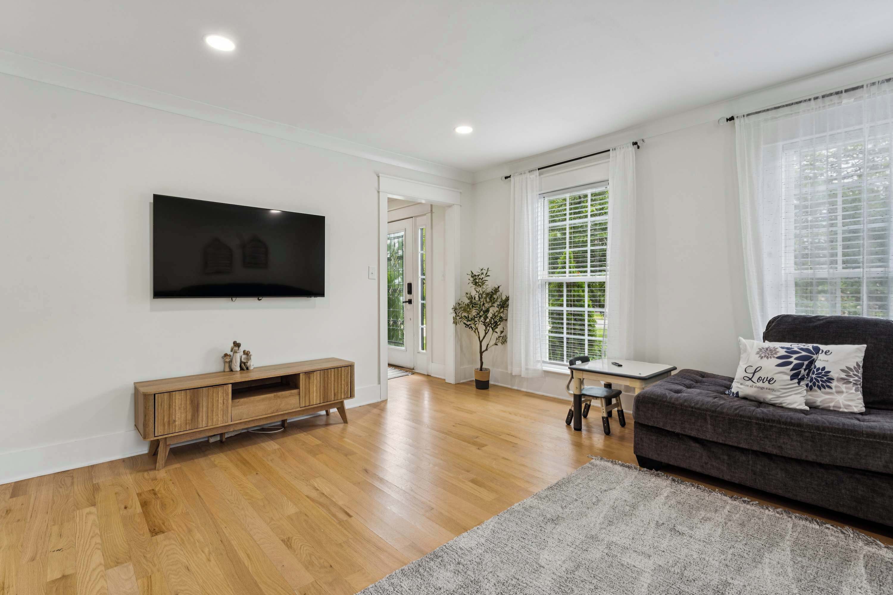 Ein Wohnzimmer mit Flachbildfernseher, Holzboden, einem dunkelgrauen Sofa, einem kleinen Tisch und einem Blumentopf.