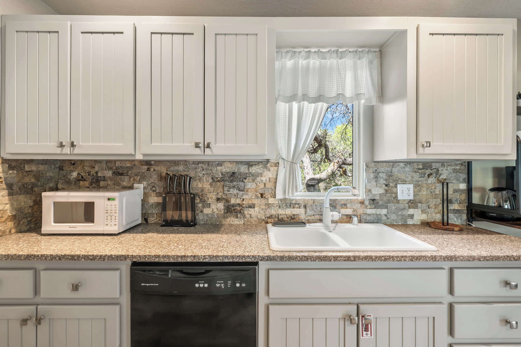 A kitchen with white cabinets, a granite countertop, a microwave, a dishwasher, and a window with a view of greenery.