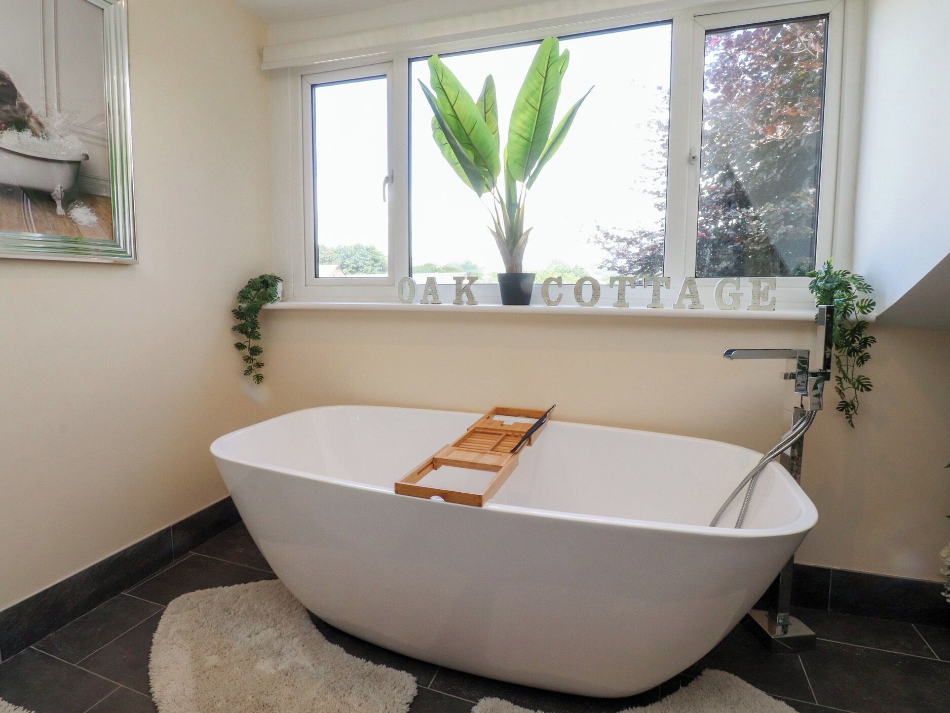Una bañera independiente con una bandeja de madera, una ventana con una planta y las palabras 'OAK COTTAGE' en el alféizar.