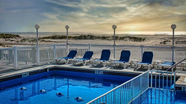 Piscina con vista a la playa y al océano, rodeada de vallas blancas y sillas de descanso azules.