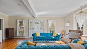 A spacious living room with a large blue sofa, a wooden floor, and a chandelier. - 8BR Luxury Farmhouse · Waterfall · Hot Tub · Catskills (Ellenville)