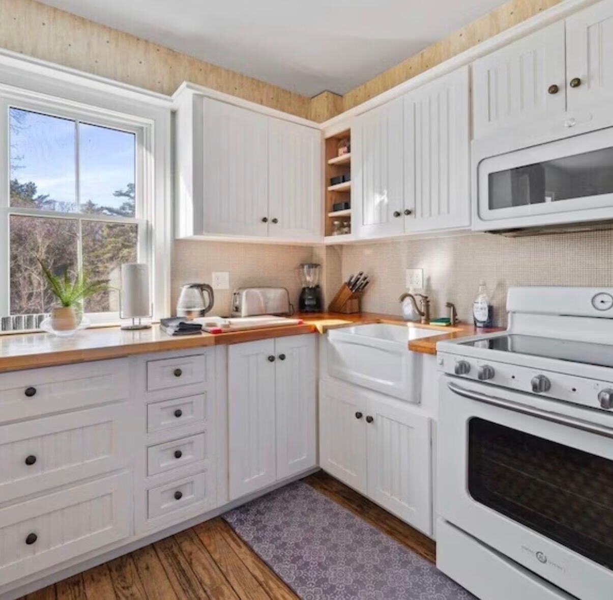 Une cuisine avec des armoires blanches, un plan de travail en bois, une cuisinière, un four à micro-ondes et une fenêtre donnant sur les arbres.