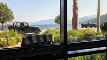 A view from inside a room with a window overlooking a beach with parked vehicles and a large tree.