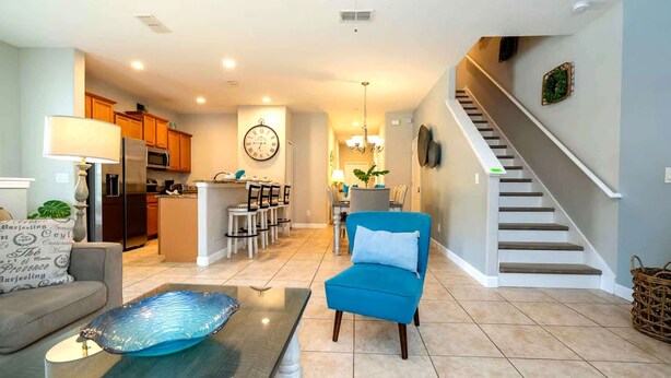 A modern living room with a sofa, a blue armchair, a glass coffee table, a kitchen in the background, and a staircase. - Golden Hours at Paradise Palms (Kissimmee)
