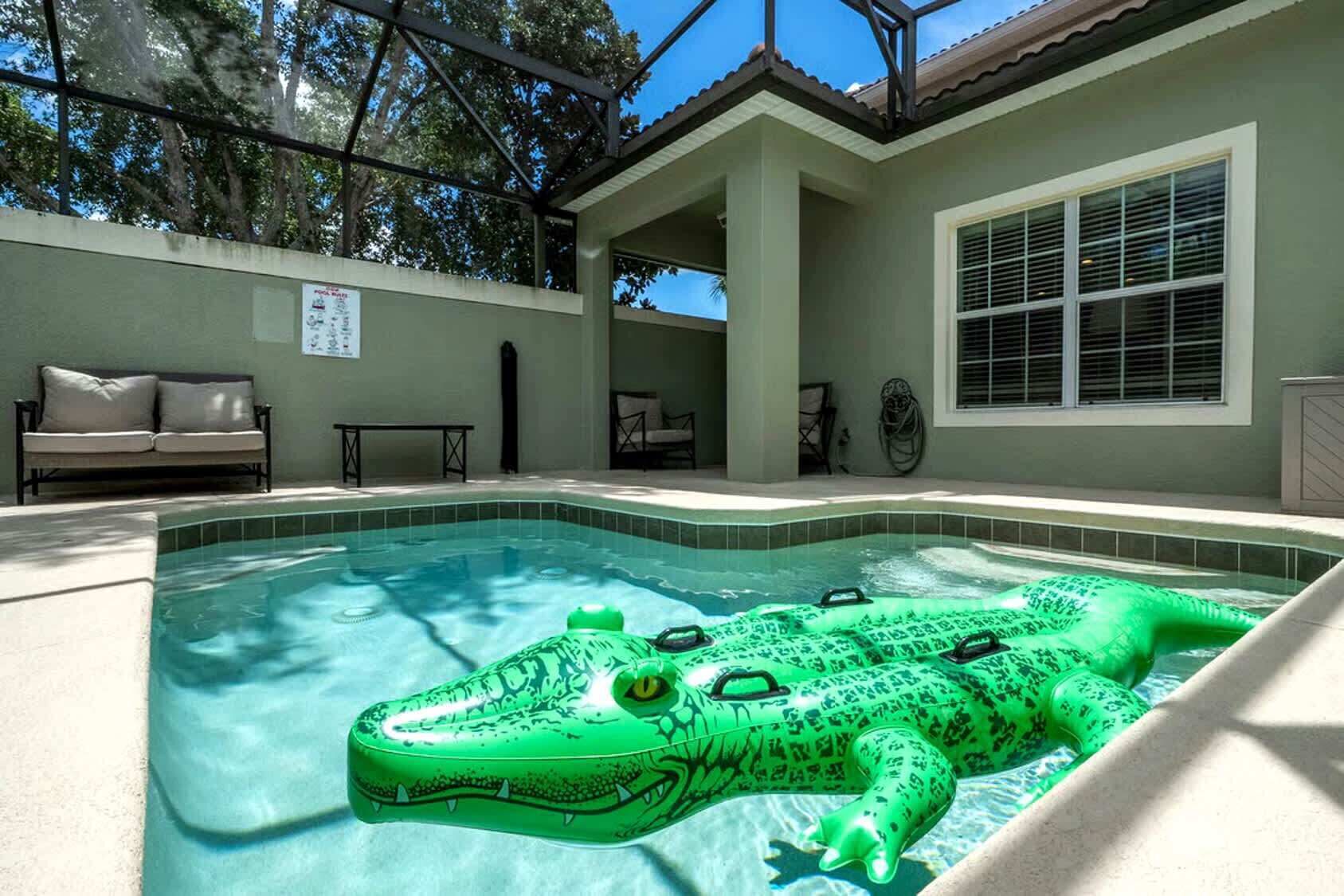 A swimming pool with a green inflatable alligator, a covered patio area, and a beige sofa.