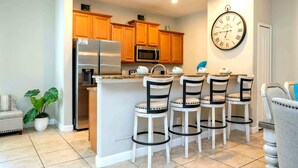 A modern kitchen with wooden cabinets, a stainless steel refrigerator, a microwave, and a bar area with four stools. - Golden Hours at Paradise Palms (Kissimmee)