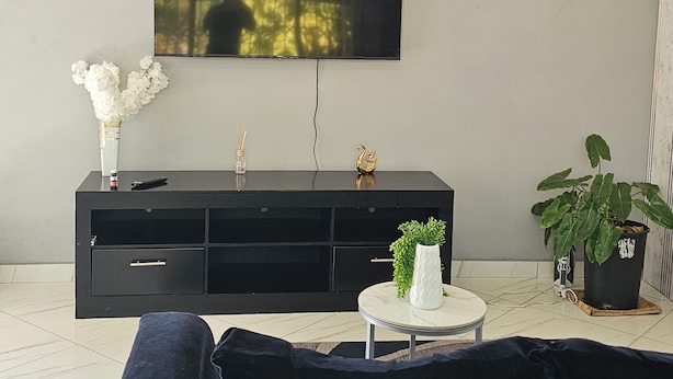 Una sala de estar con un mueble multimedia negro, un televisor de pantalla plana montado en la pared, un sofá oscuro y una mesita redonda con una planta.