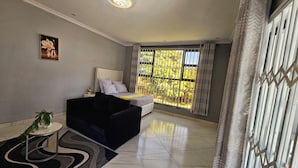 Un dormitorio moderno con una cama, una mesita con una planta, un adorno en la pared y un ventanal con vistas a zonas verdes.