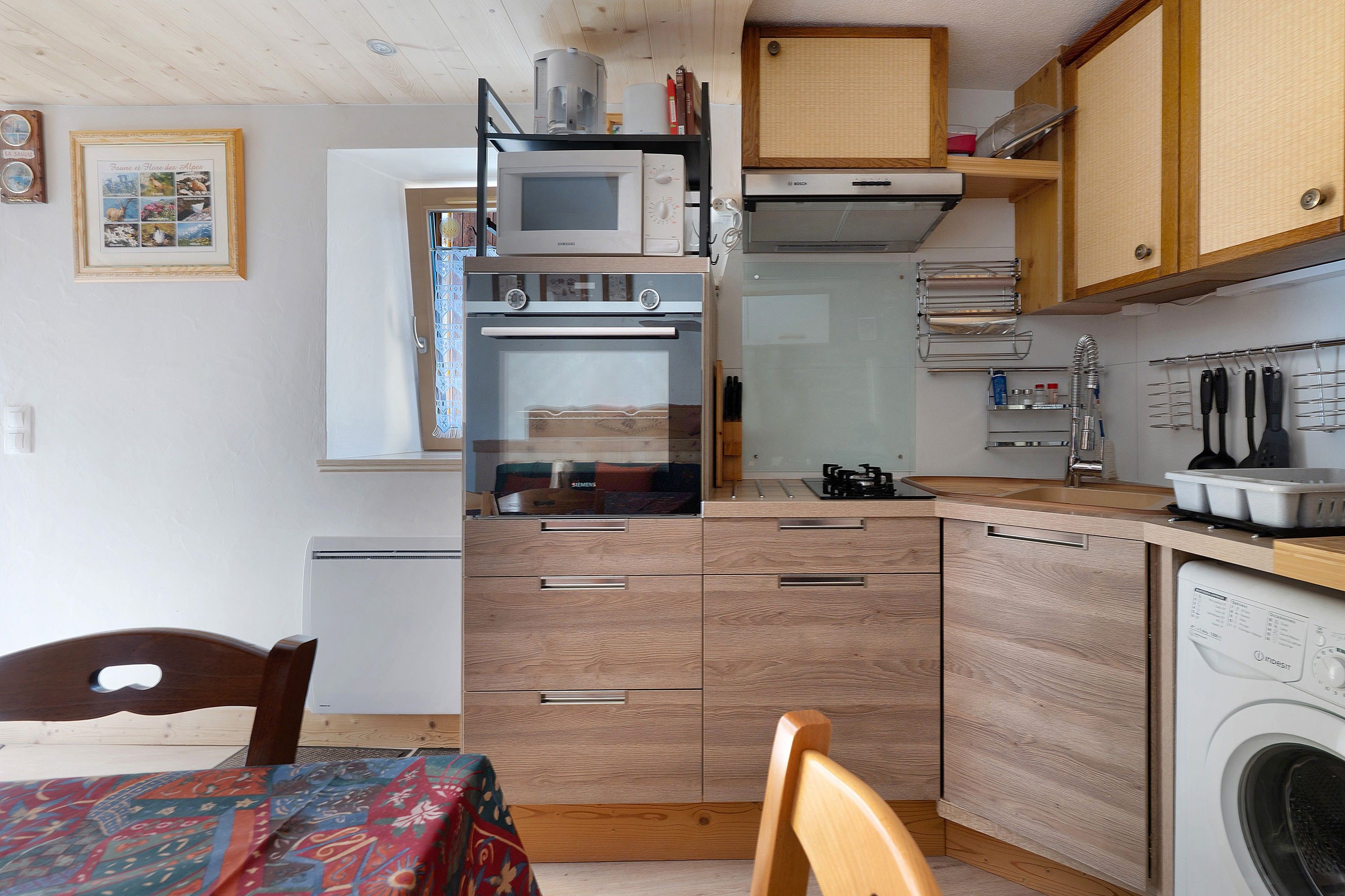 Une cuisine compacte équipée d’un four à micro-ondes, d’un four et d’un lave-vaisselle. On y trouve une table à manger avec des chaises et une nappe à motifs.
