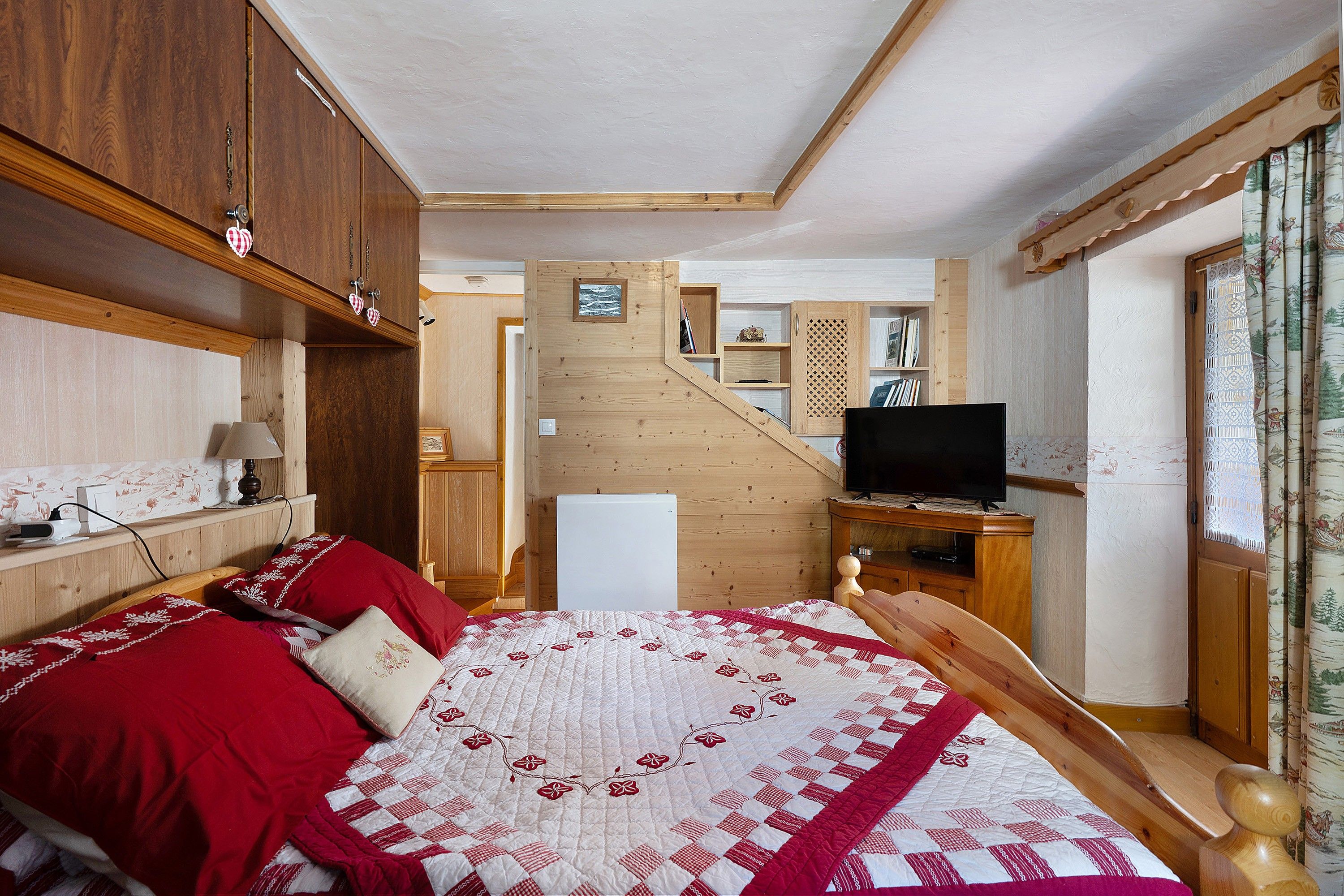 A bedroom with a bed, wooden wardrobe, television, and a small table with a lamp.