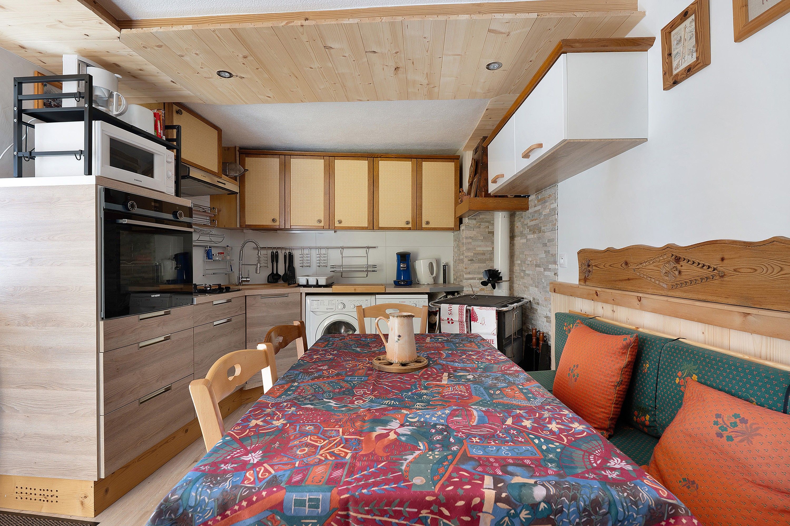 A compact kitchen with wooden cabinets, a microwave, a washing machine, and a dining area with a patterned tablecloth.