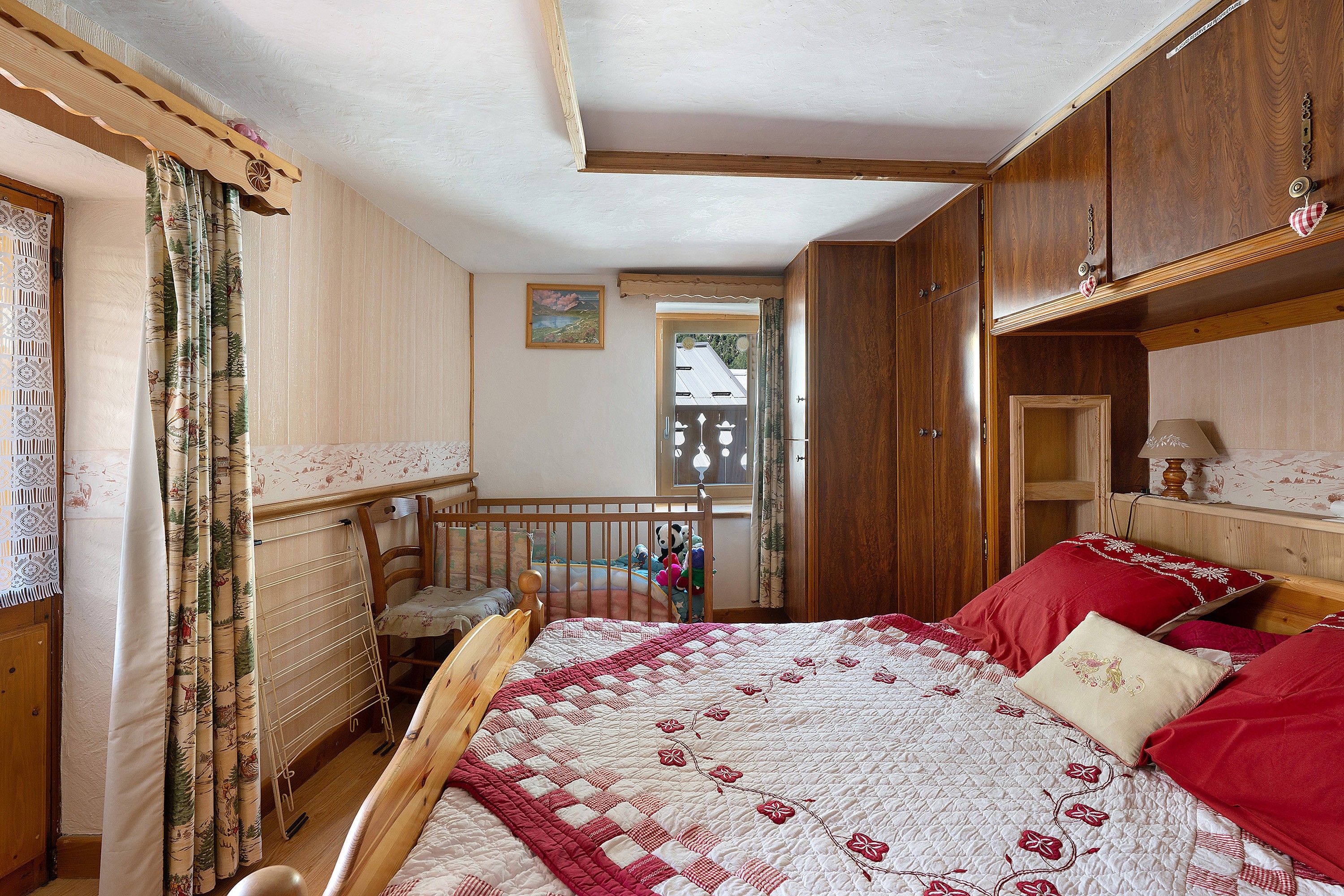 Une chambre à coucher avec un lit en bois, une chaise en bois, une fenêtre donnant sur un paysage agréable et une porte menant à une autre pièce.