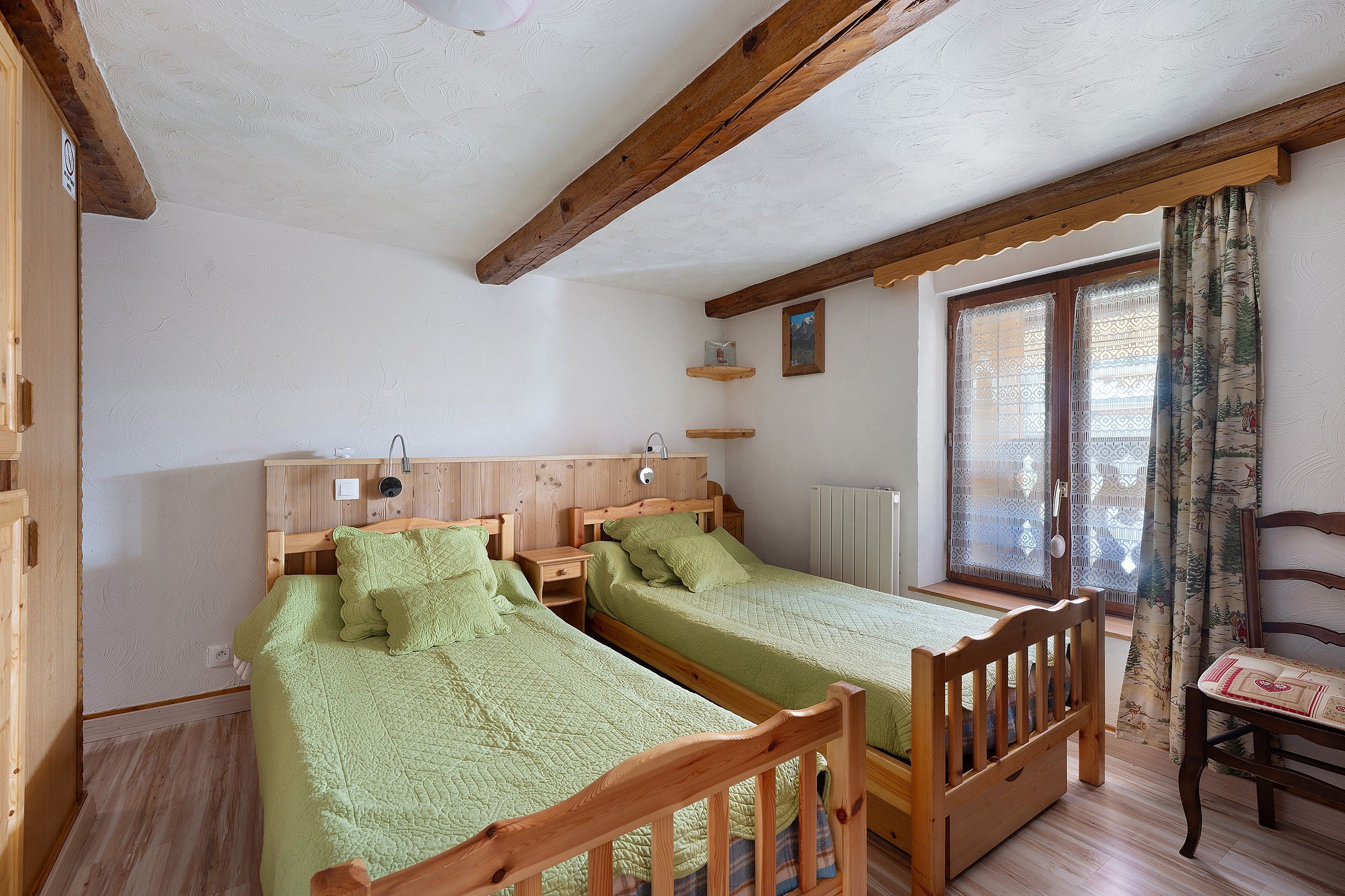 A room with two wooden beds, a wooden chair, a window with curtains, and a radiator.