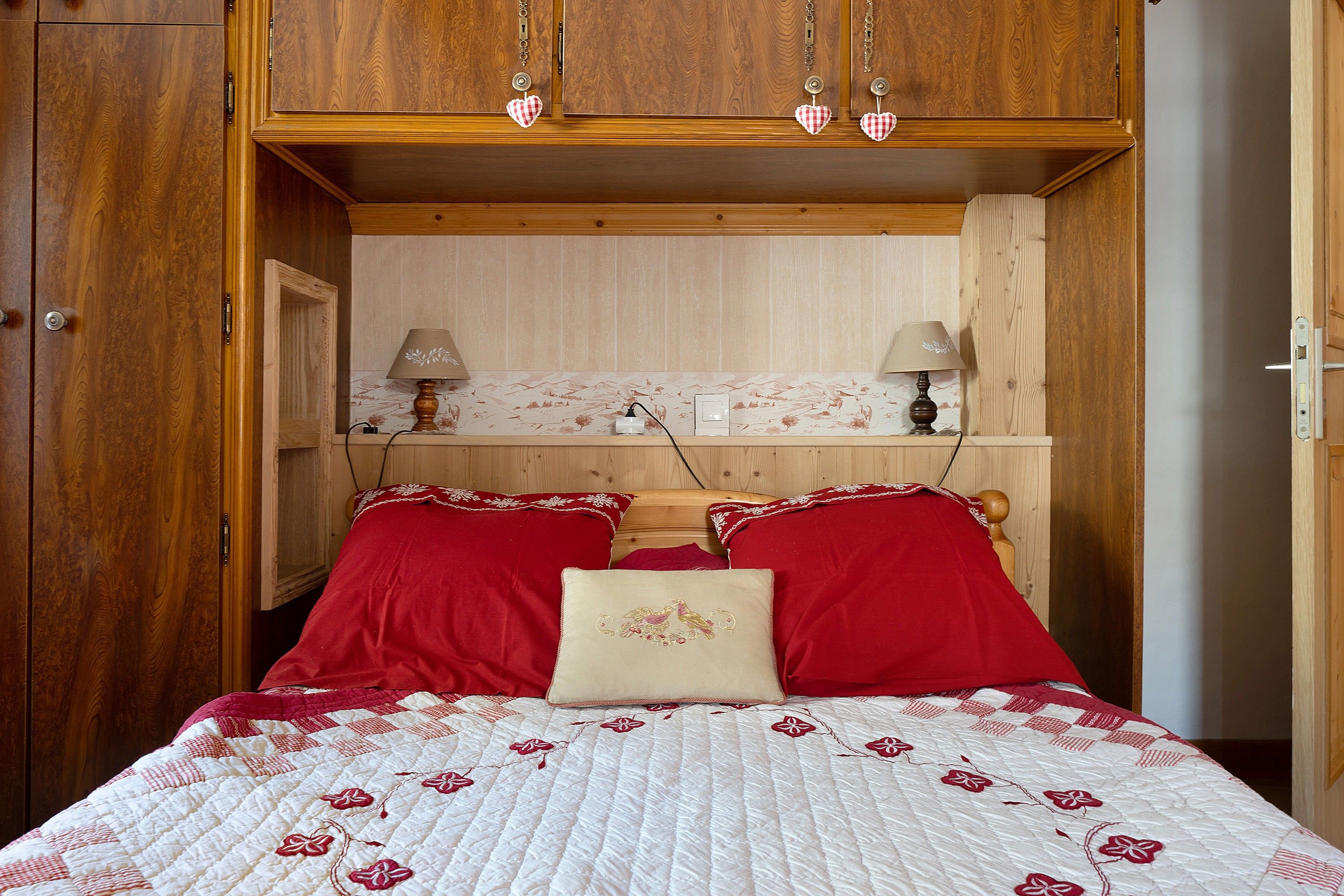 A bedroom with a wooden wardrobe, a bed with red and white bedding, two lamps on a shelf, and a wall with a floral wallpaper.