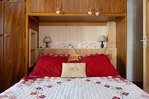 Ein Schlafzimmer mit einem Holzschrank, einem Bett mit rot-weißer Bettwäsche, zwei Lampen auf einem Regal und einer Wand mit floralem Wandpapier.