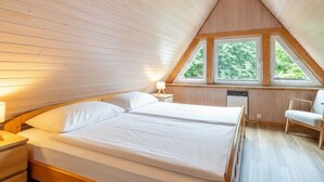 Makuuhuoneessa on puinen katto, sänky, yöpöytä, tuoli ja ikkuna, josta avautuu näkymä puihin.