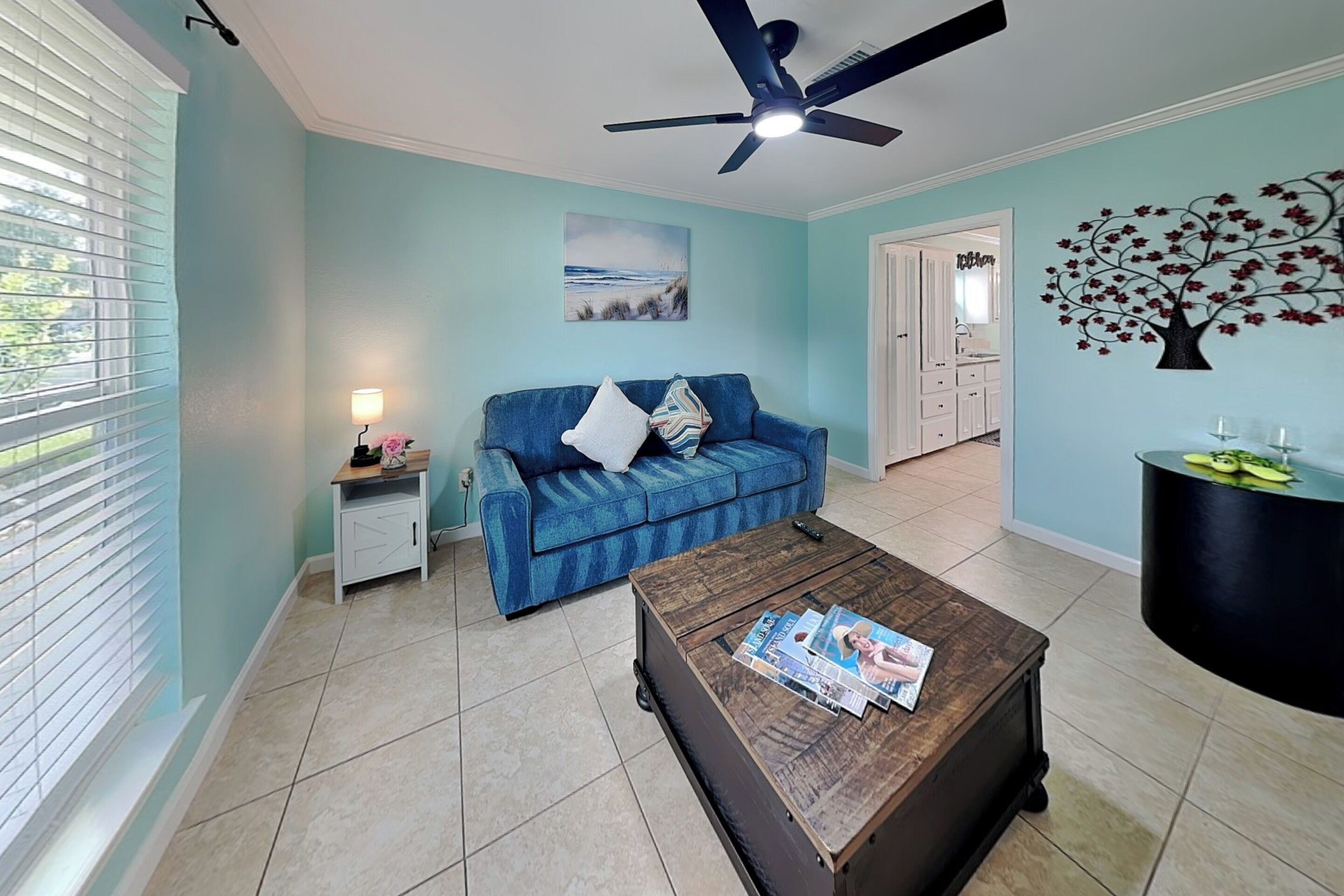 A living room with a blue sofa, a coffee table with magazines, a ceiling fan, and a wall with a tree decoration.