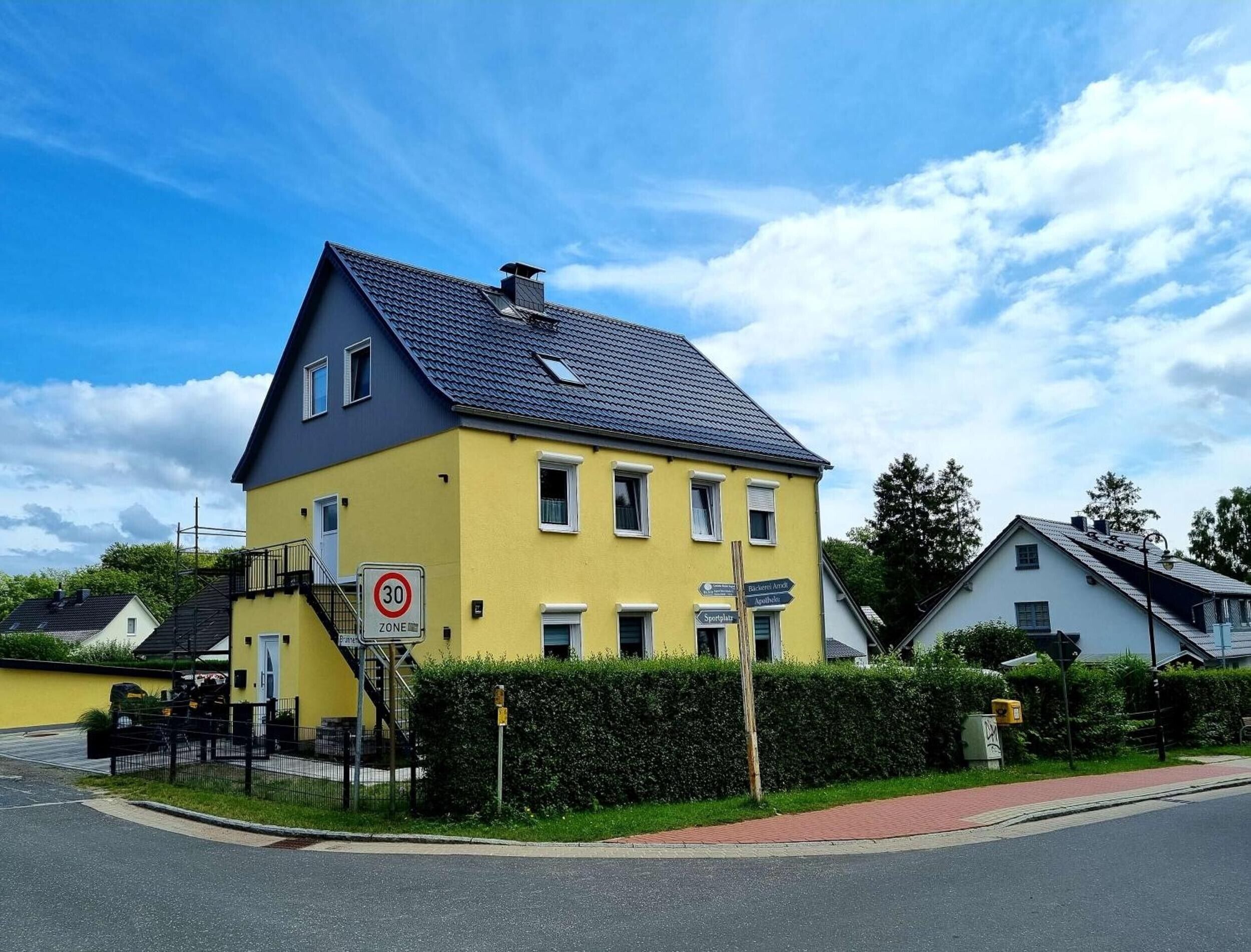 Ein gelbes, zweistöckiges Gebäude mit dunklem Dach und einer sichtbaren Treppe, sowie einem Geschwindigkeitsbegrenzungsschild.