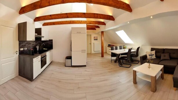 Un moderno loft con cucina, zona pranzo e soggiorno, caratterizzato da travi a vista in legno, una TV a schermo piatto e un'ampia finestra.