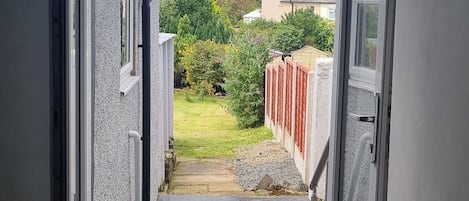 A narrow pathway leading to a garden with a red fence, a small pond, and a house in the background.