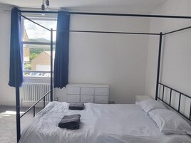 A four-poster bed with white linens, a radiator, and a window with blue curtains.