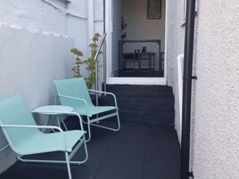Un pequeño espacio al aire libre con dos sillas azul claro y una mesa, una planta en maceta y vistas de un edificio con una escalera que conduce a un balcón.