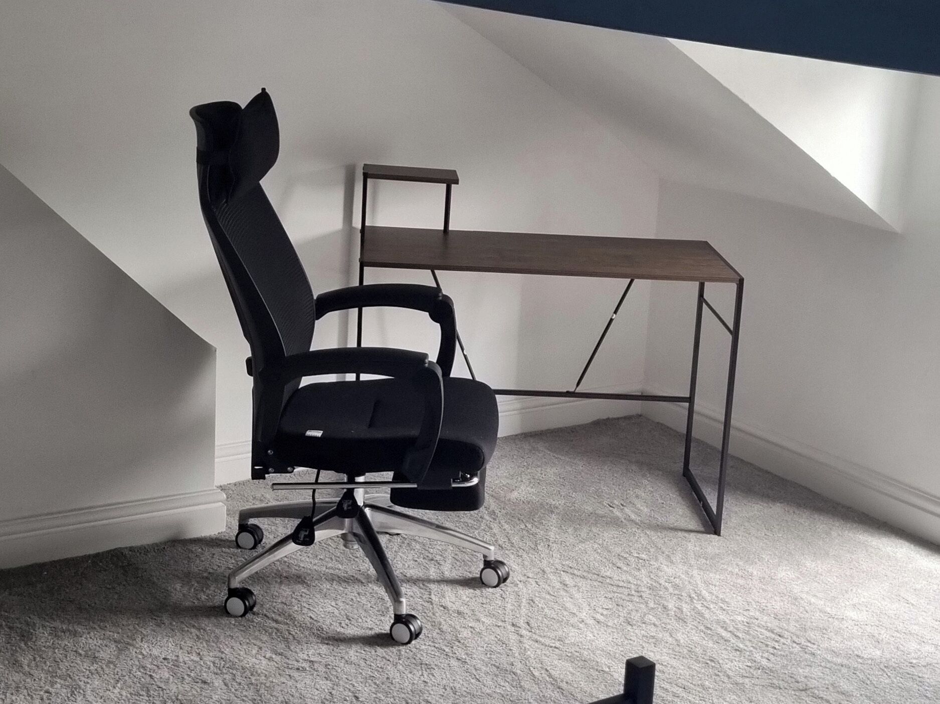 Une chaise de bureau noire à roulettes, un bureau en bois et un plafond en pente dans une pièce dont le sol est recouvert de moquette.