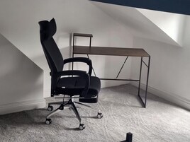 Een zwarte bureaustoel met wielen, een houten bureau en een schuin dak in een kamer met vloerbedekking.