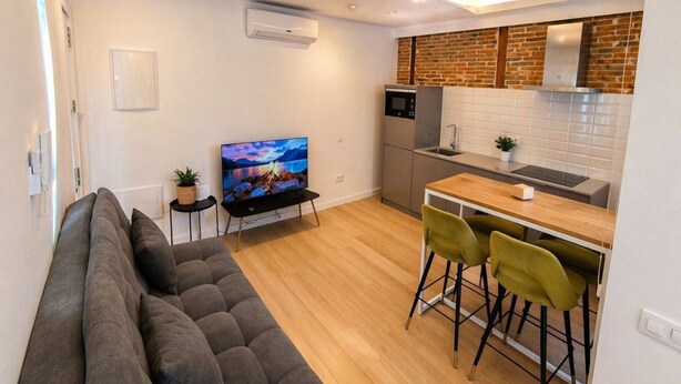 A modern living room with a sofa, a television, and a kitchen area with a brick wall and wooden beams. - Stay Near El Rastro for 4 (Madrid)