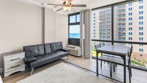 A modern living room with a black leather sofa, a dining table with chairs, a ceiling fan, and a large window with a view of a high-rise building. - 🌴Amazing Ocean Views | Beachfront Pool | Oceanside Inn 605🌴 (Daytona Beach)