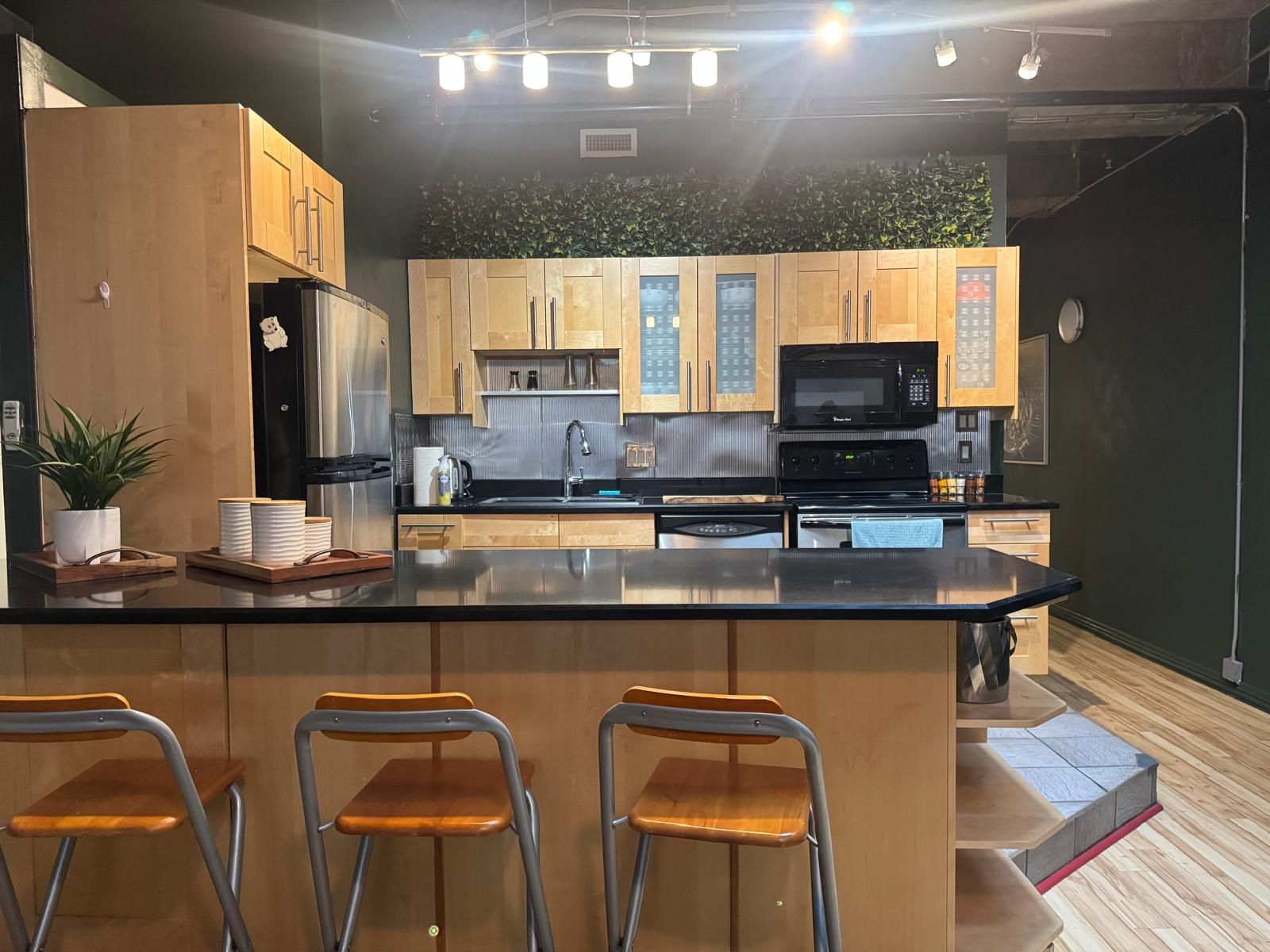 Una cucina moderna con mobili in legno, un piano di lavoro nero, elettrodomestici in acciaio inossidabile e tre sgabelli da bar.