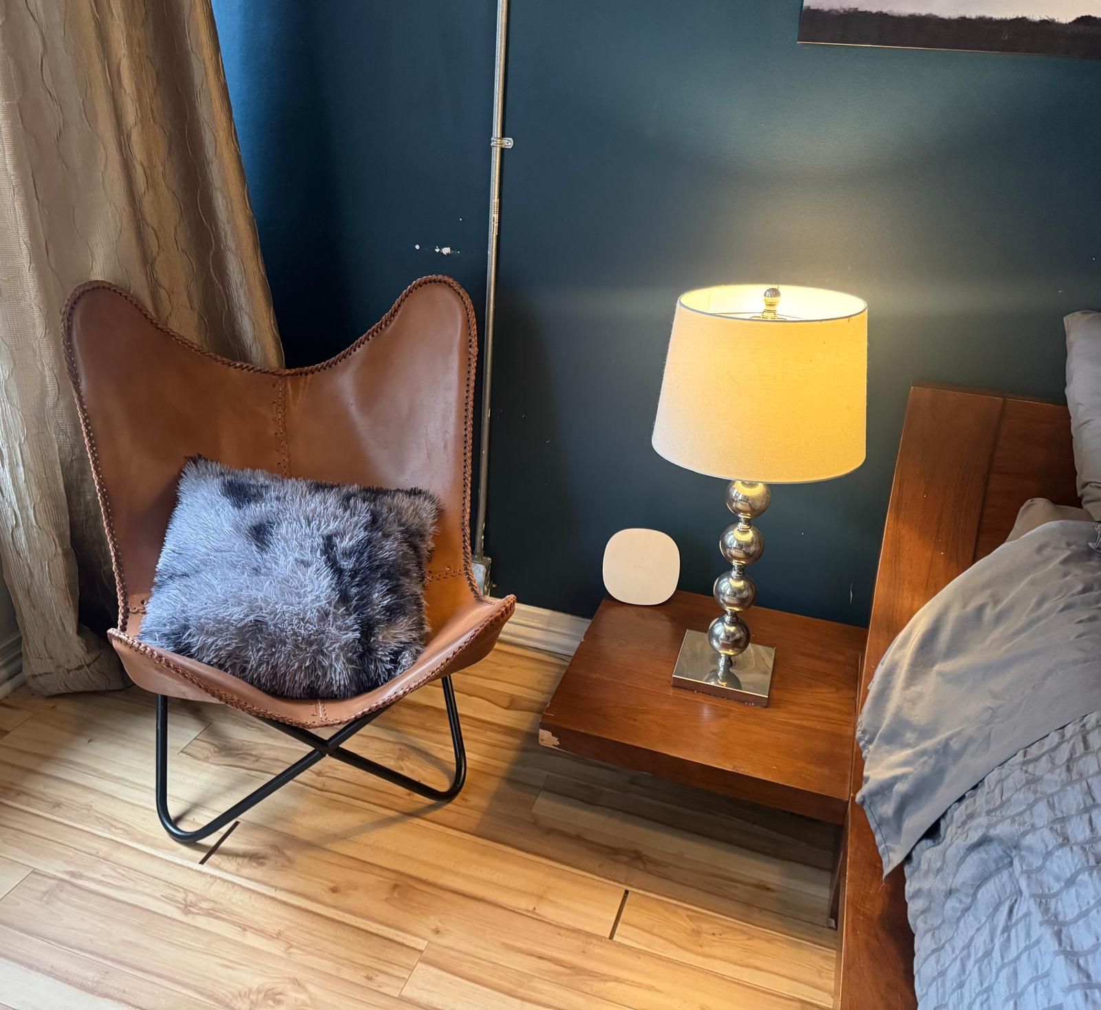 Une pièce avec un fauteuil en cuir marron, un coussin moelleux, une table de chevet en bois avec une lampe et un lit recouvert d’une couverture grise.