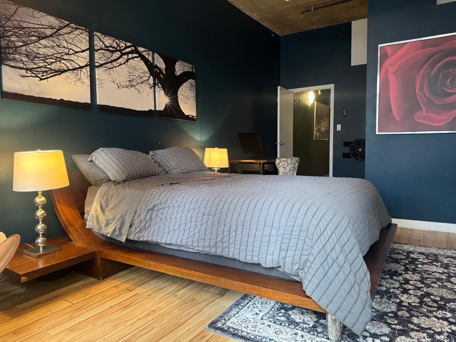 A bedroom with a bed, two bedside lamps, a large wall art of a tree, a door, and a patterned rug.