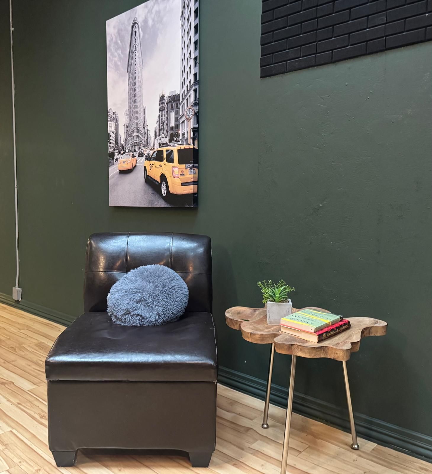Ein Zimmer mit einem schwarzen Ledersessel, einem kleinen Holfnachtkästchen mit Büchern und einer Pflanze sowie einem großen Stadtbild an der Wand.