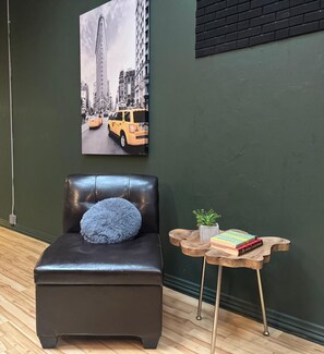 Une pièce comprenant un fauteuil en cuir noir, une petite table d’appoint en bois avec des livres et une plante, et un grand tableau représentant un paysage urbain accroché au mur.