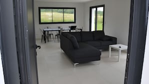 A modern living room with a sectional sofa, a dining table with chairs, and a large window offering a view of a field. - Villa Pradel – Holiday Home Guérande La Baule, 4 Bedrooms, 7 Guests (Guérande)