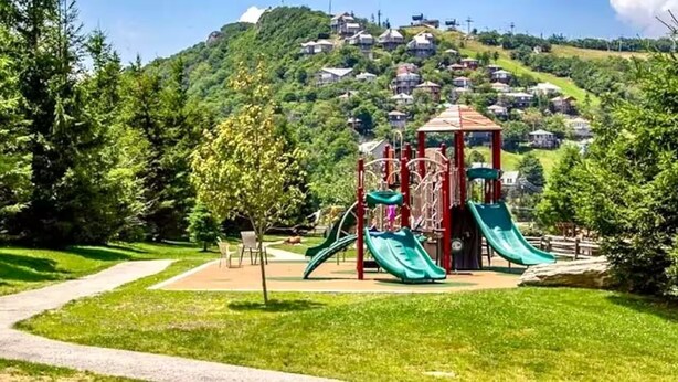 Ein Spielplatz mit Rutschen und Klettergerüsten, umgeben von Bäumen und einem Weg zu einem Wohngebiet.